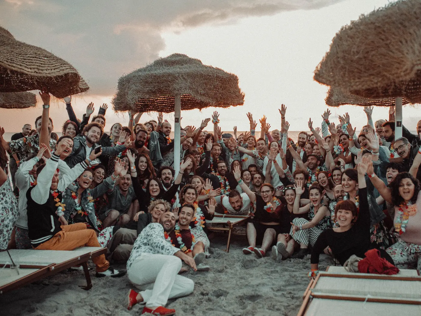 Beach party at sunset: everyone together, arms in the air, among the straw umbrellas
